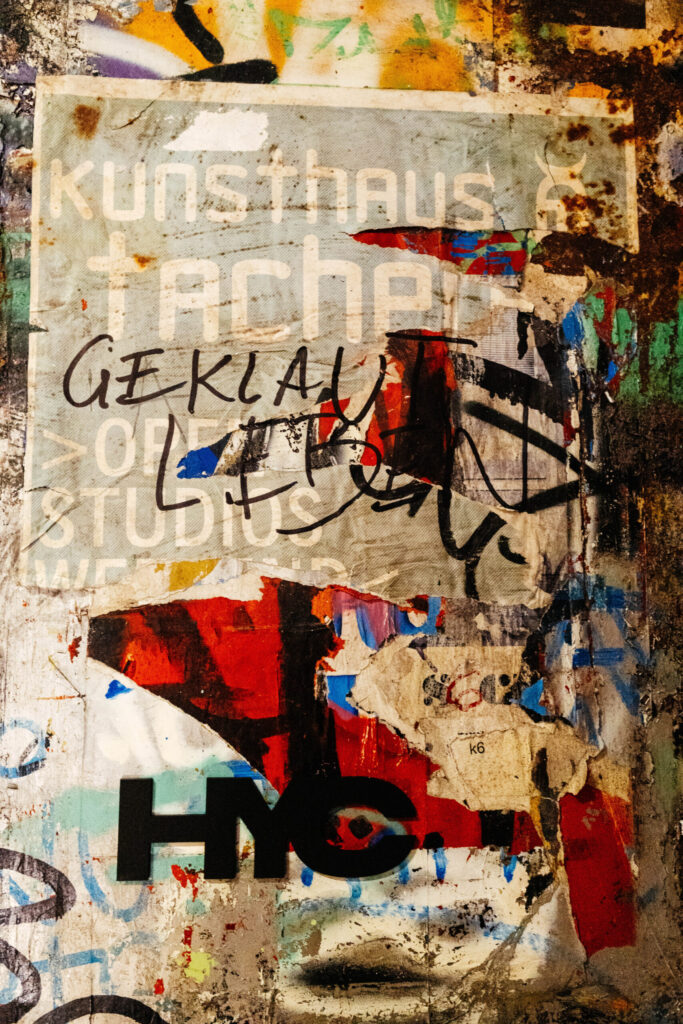 Plakatfragment mit der Aufschrift „kunsthaus tacheles“ auf einer von Aufklebern, Farbschichten und Kritzeleien überlagerten Wand. Über das Plakat wurde mit schwarzem Marker das Wort „GEKLAUT“ geschrieben. Darunter sind weitere Schriftzüge, teils unleserlich, sowie bunte Reste von Papier, Farbe und Tags. Am unteren Rand ist das schwarze Kürzel „HYC“ zu sehen. Die Collage wirkt wie ein visuelles Manifest des Widerstands – rau, laut und chaotisch – ganz im Stil der alternativen Kunstszene des ehemaligen Berliner Kunsthauses Tacheles.