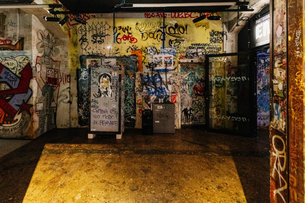 Innenraum mit intensiv besprühten Wänden, Türen und Objekten im Stil eines urbanen Kunstzentrums. Die gesamte Fläche ist mit Graffiti, Tags, Stickern und Wandzeichnungen bedeckt – darunter politische Botschaften, Portraits, Schriftzüge und bunte Kritzeleien. Links ist ein großes Wandbild mit der Aufschrift „STOP“ zu sehen, rechts ein Glaseingang mit dem Schriftzug „PHOTOGRAPHY ENTRANCE“. In der Mitte stehen zwei mit Graffiti überzogene Kästen oder Schränke. Die Szene wirkt chaotisch, kreativ und rebellisch – ein typisches Merkmal der Kunstszene im ehemaligen Berliner Tacheles.
