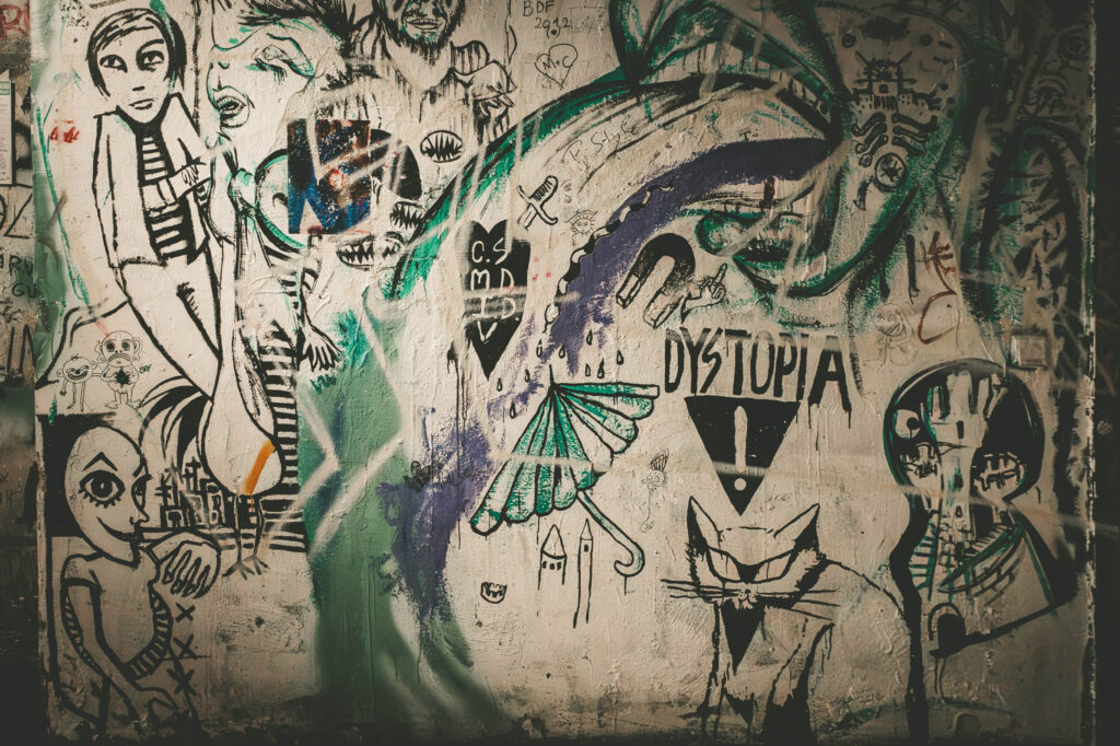 Bunte Wand voller Streetart und Kritzeleien im Stil eines künstlerischen Chaos. Die Wand zeigt zahlreiche schwarz-weiße Zeichnungen von Figuren mit übergroßen Augen, Fratzen, einer Katze im Anzug, einem Schirm mit Regentropfen, Symbolen wie einem Magneten und dem Wort „DYSTOPIA“ mit Ausrufezeichen. Grüne und violette Farbschlieren sowie Graffiti-Tags überlagern die Illustrationen. Die Szene wirkt wie ein visuelles Tagebuch urbaner Gedankenwelt – expressiv, anarchisch und voller Symbolik, typisch für das ehemalige Berliner Kunsthaus Tacheles.