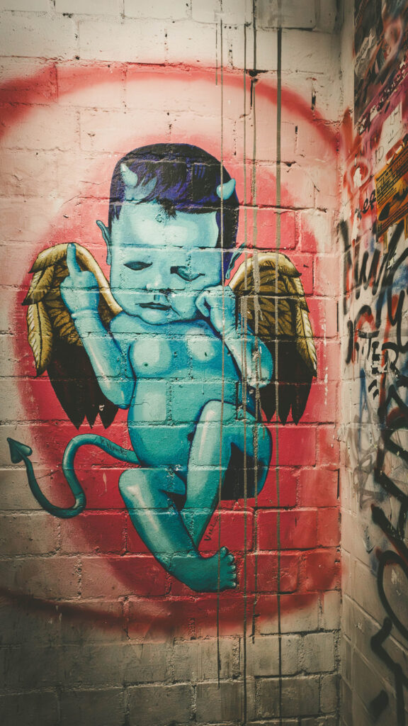 Streetart eines blauhäutigen, schlafenden Teufelsbabys mit kleinen Hörnern, goldenen Engelsflügeln und spitzem Teufelsschwanz, gemalt auf eine Backsteinwand. Das Baby zeigt provokativ den Mittelfinger. Der Hintergrund ist in Rosa und Rot gesprüht, wodurch sich die Figur farblich stark abhebt. Rechts neben dem Bild sind weitere Graffiti und Posterreste zu erkennen. Das Werk kombiniert kindliche Unschuld mit rebellischen Elementen – typisch für die subversive Ästhetik des ehemaligen Berliner Kunsthauses Tacheles.