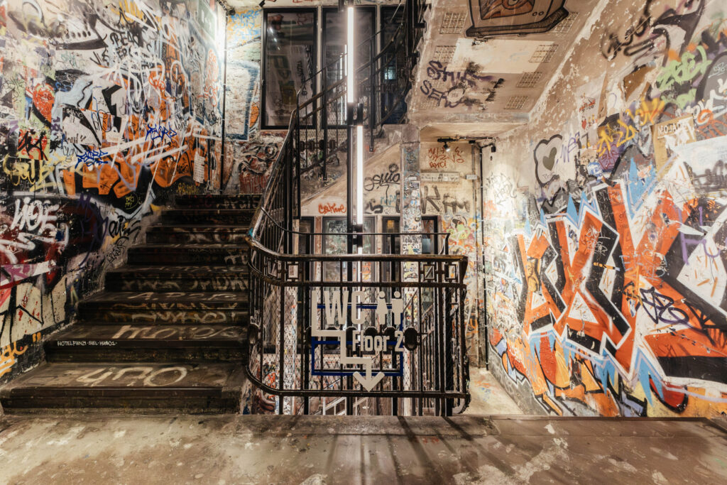 Innenaufnahme eines bunt besprühten Treppenhauses mit intensiven Graffiti an Wänden, Decke und Treppenstufen. Ein schwarzes Geländer umgibt das Treppenauge, an dem ein Schild mit der Aufschrift „WC Floor 2“ und Piktogrammen für Toiletten angebracht ist. Eine vertikale Leuchtstoffröhre verläuft mittig durch das Bild und beleuchtet den Raum. Die Atmosphäre ist kreativ, chaotisch und urban – ein Sinnbild für das alternative Flair des ehemaligen Kunsthauses Tacheles in Berlin.