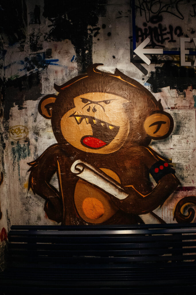 Bunte Streetart eines comicartig dargestellten, grimmig dreinblickenden Affen an einer mit Graffiti übersäten Wand. Der Affe hat ein rotes Maul mit spitzen Zähnen, hält eine zusammengerollte Schriftrolle in der Hand und trägt eine schwarze Armbinde mit roten Punkten. Im oberen rechten Bildbereich weist ein weißer Pfeil nach links. Vor dem Wandbild steht eine dunkle Metallbank. Die Szene ist schwach beleuchtet und wirkt urban und roh.