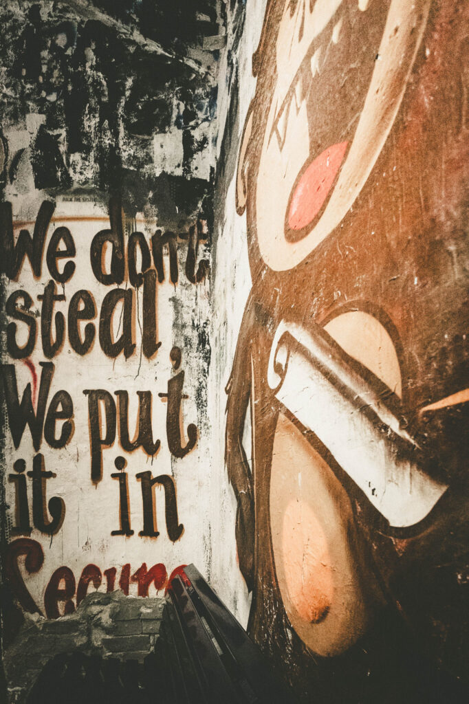 Wandgraffiti mit einem großflächigen, comicartigen Affen, der eine Schriftrolle in der Hand hält und wütend den Mund geöffnet hat. Neben dem Affen steht in brauner und roter Schrift der ironisch-provokante Text: „We don’t steal, we put it in secure“. Die Wand ist stark abgenutzt, mit zahlreichen Farbschichten, Kritzeleien und Textfragmenten. Am unteren Bildrand sind schwarze Sitzbänke zu erkennen. Die Szene vermittelt eine rebellische, subkulturelle Atmosphäre – typisch für das ehemalige Berliner Kunsthaus Tacheles.
