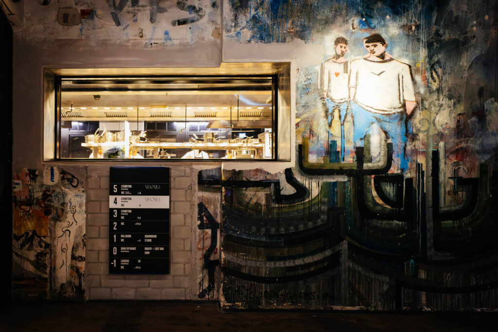 Wandkunst in einem urbanen Innenraum mit einem großen Fenster, durch das eine moderne Restaurantküche sichtbar ist. Links unter dem Fenster befindet sich ein schwarzes Orientierungsschild mit Etagenangaben und dem Namen „Verniki“. Die Wand ist mit farbenfrohen, abstrakten Graffiti und einem Wandgemälde zweier Männer in weiß-blauen Outfits bemalt. Einer der Männer hat ein rotes Herz auf der Brust. Die Wandgestaltung ist vielfältig und detailreich – typisch für das ehemalige Kunsthaus Tacheles in Berlin, das heute Teile von „Fotografiska“ beherbergt.