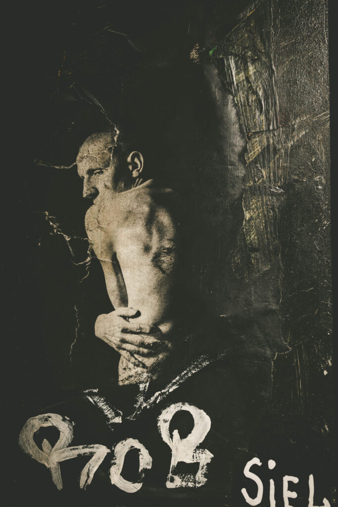 Kunstvoll gestaltetes Schwarz-Weiß-Plakat eines nackten Mannes, der mit dem Rücken zur Kamera steht und sich schützend die Arme um den Oberkörper legt. Der Körper wirkt angespannt und verletzlich. Die Oberfläche des Posters ist rissig und abgenutzt, was dem Bild eine raue, verwitterte Ästhetik verleiht. Unten sind großflächige weiße, teils unleserliche Buchstaben zu sehen, darunter das Wort „SIEL“. Die Szene ist düster beleuchtet und wirkt emotional intensiv, fast intim – typisch für die expressive Kunst im ehemaligen Kunsthaus Tacheles in Berlin.