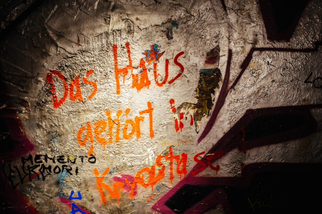 Graffiti auf einer rau verputzten Wand mit dem handschriftlich gesprühten Text in Orange und Rot: „Das Haus gehört in Künstlerhand“. Der Schriftzug ist leicht verwischt und von weiteren Tags, Kritzeleien und Farbspritzern umgeben. Links unten steht in schwarzer Schrift „Memento Mori“, daneben kleine Botschaften wie „stay alive“. Die Wand ist bunt, wild und ausdrucksstark – ein klares politisches Statement zur kulturellen Nutzung des Hauses, typisch für das ehemalige Kunsthaus Tacheles in Berlin.