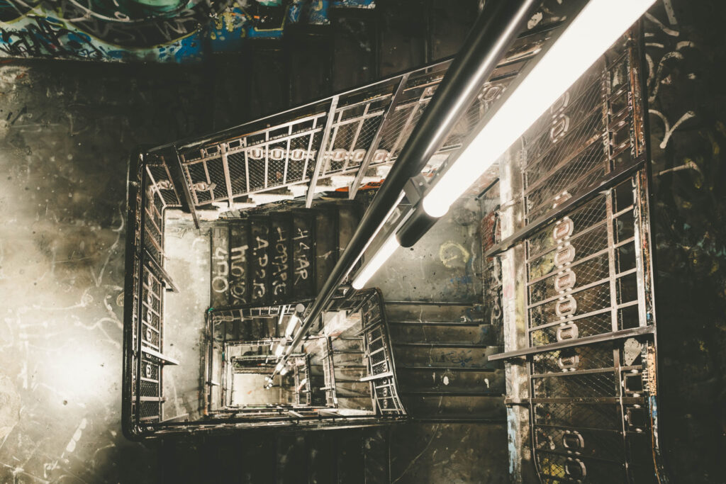 Blick in ein enges, grafisch anmutendes Treppenhaus mit Metallgeländern, Gittern und zentral verlaufenden Neonröhren. Die Perspektive zeigt den Schacht spiralförmig nach unten, wodurch ein visueller Sog entsteht. Die Wände und Treppenstufen sind mit Graffiti, Tags und Kritzeleien bedeckt. Die Beleuchtung betont die industrielle Atmosphäre, während die abgenutzten Strukturen und Farbschichten den rauen Charme des ehemaligen Kunsthauses Tacheles in Berlin widerspiegeln.