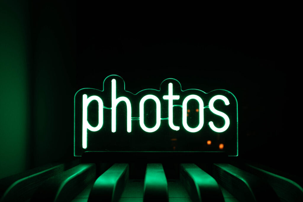 Leuchtendes, grünliches Neon-Schild mit dem Wort „photos“ in einer klaren, modernen Schriftart. Das Schild steht auf einer dunklen Oberfläche mit gebogenen Streben oder Holzlatten, die im Vordergrund sichtbar sind. Der Hintergrund ist schwarz, wodurch das grüne Licht besonders stark hervorsticht. Die Szene wirkt stilvoll, minimalistisch und erinnert an eine moderne Fotoausstellung oder ein Fotostudio. Es setht im Kontext von Fotografiska Berlin.