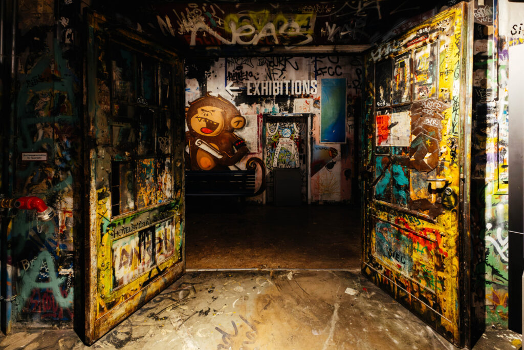 Blick durch zwei weit geöffnete, bunt besprühte Metalltüren in einen düsteren Innenraum voller Graffiti. An der Rückwand ist ein großes Wandbild eines comicartigen Affen mit wütendem Gesichtsausdruck und einer Schriftrolle in der Hand zu sehen. Darüber weist der Schriftzug „→ EXHIBITIONS“ den Weg. Die gesamte Szene ist von einem urbanen, künstlerischen Chaos geprägt, mit farbintensiven Graffiti, Stickern und Kritzeleien – typisch für das ehemalige Berliner Kunsthaus Tacheles.
