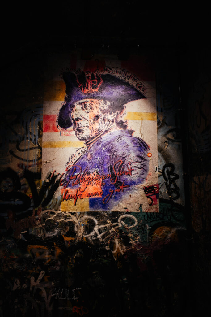 Streetart-Porträt eines historischen Mannes mit Dreispitz und Feder, vermutlich Friedrich der Große, auf eine stark abgenutzte Wand gemalt. Das Bild ist in Blau-, Rot- und Gelbtönen gehalten, mit einer markanten kalligrafischen Inschrift in Rot: „Die Religionen sind...“ (vermutlich ein Zitat von Friedrich dem Großen). Das Porträt hebt sich kontrastreich vom von Graffiti und Tags überzogenen Hintergrund ab. Die Szene ist dramatisch beleuchtet, was die Textur der Wand und das Kunstwerk hervorhebt. Der Ort wirkt wie ein Relikt alternativer Berliner Subkultur.