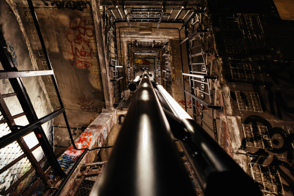 Blick nach oben durch ein mehrstöckiges, enges Treppenhaus mit industriellem Charme. Der Blick wird von einem mittig verlaufenden Rohr oder einer Stange in die Tiefe gezogen, die sich perspektivisch verjüngt. Die Wände und Geländer des Treppenhauses sind stark mit Graffiti, Tags und Abnutzungsspuren bedeckt. Metallgitter, rostige Elemente und Beton bestimmen die Szenerie. Die Beleuchtung ist schummrig, mit gezielten Lichtreflexen auf dem Rohr. Die Atmosphäre wirkt roh, urban und leicht klaustrophobisch – typisch für das ehemalige Kunsthaus Tacheles in Berlin.