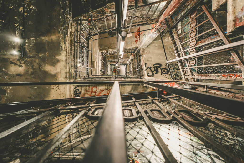 Blick in das Innere eines mehrstöckigen, verlassen wirkenden Treppenhauses mit industriellem Charakter. Die Perspektive zeigt den vertikalen Schacht von unten nach oben, dominiert von Metallgittern, Rohren und einer zentralen Lichtleiste. Die Wände sind mit Graffiti bedeckt und stark abgenutzt, was dem Ort einen urbanen, rauen Charme verleiht. Metallische Strukturen und Gitter fangen das Licht ein und erzeugen geometrische Muster. Die Szene wirkt verlassen, geheimnisvoll und erinnert an das ehemalige Berliner Kunsthaus Tacheles.