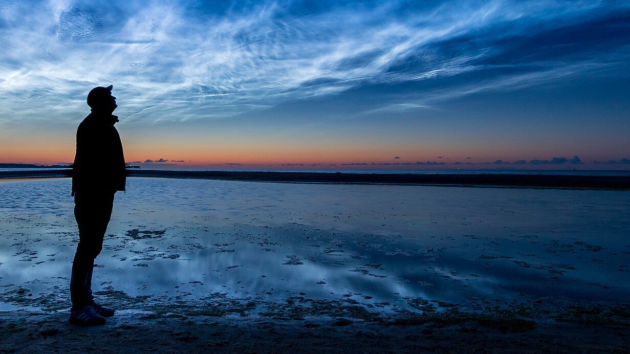 Silhouette eines Mannes am Strand bei Sonnenuntergang, der in den Himmel mit beeindruckenden Wolkenformationen blickt. Der ruhige Wasserspiegel reflektiert die Farben des Himmels.