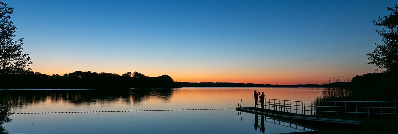 Paar steht am Ufer eines ruhigen Sees bei Sonnenuntergang, umgeben von Bäumen und einer malerischen Landschaft.