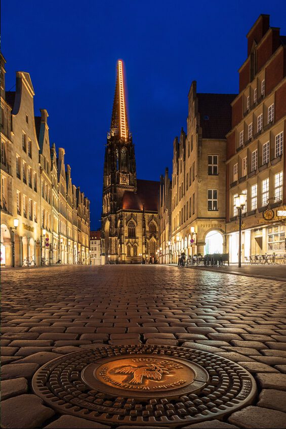 Nachtaufnahme des Münsteraner Prinzipalmarkts mit der St. Lamberti-Kirche im Hintergrund, beleuchtet von der Abenddämmerung. Die gepflasterte Straße und historische Gebäude umrahmen den Platz, während eine Münze im Vordergrund sichtbar ist.
