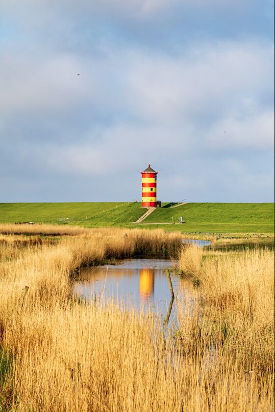 Leuchtturm in rot-gelber Streifenfarbe auf einer Wiese, umgeben von hohem Gras und einem schmalen Wasserlauf, unter einem bewölkten Himmel.