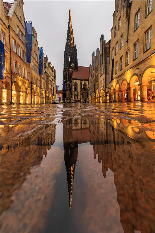 Reflexion der Lamberikirche in Münster auf nasser Pflasterstraße, umgeben von historischen Gebäuden und europäischen Flaggen, bei bewölktem Wetter.
