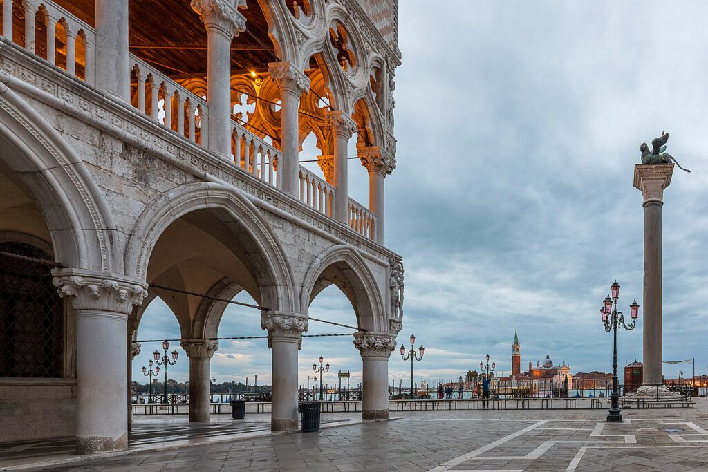 Architektonische Details des Dogenpalastes in Venedig, mit Säulen und Bögen im Vordergrund, Blick auf den Markusplatz und den Canal Grande im Hintergrund. Sanfte Beleuchtung und bewölkter Himmel schaffen eine stimmungsvolle Atmosphäre.