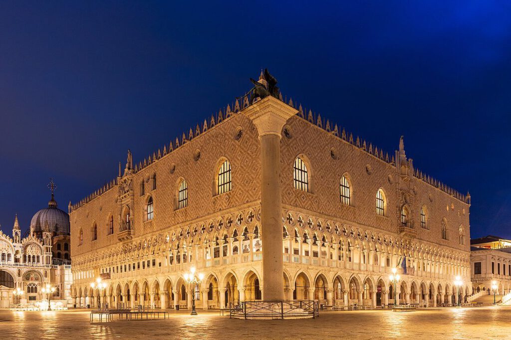 Dogenpalast in Venedig bei Nacht, beleuchtet mit warmem Licht, zeigt architektonische Details und gotische Elemente, umgeben von leeren Plätzen und historischen Gebäuden.