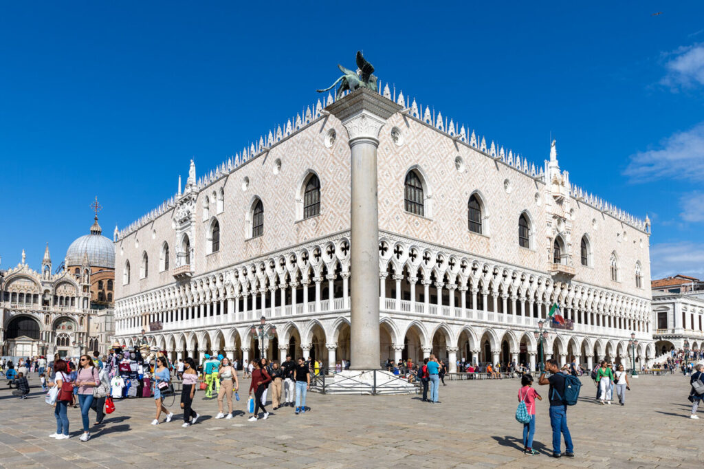 Der Markusplatz in Venedig mit dem beeindruckenden Dogenpalast, der für seine prächtigen architektonischen Details und die lebhafte Atmosphäre bekannt ist. Touristen schlendern um das Gebäude, während die Sonne am klaren blauen Himmel scheint.