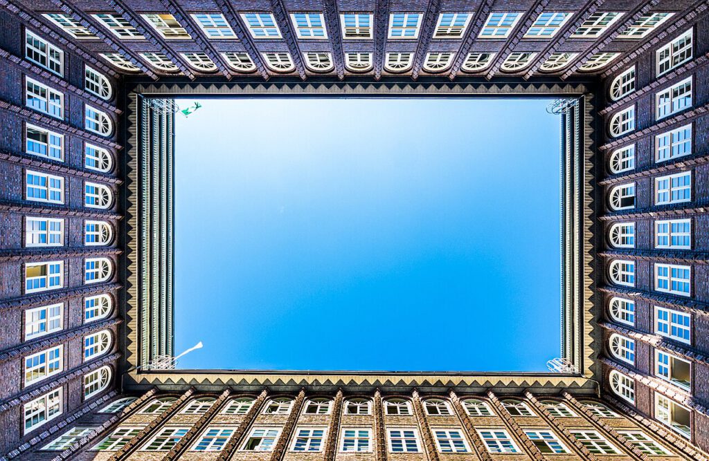Blick in den Innenhof eines historischen Gebäudes mit rotbraunen Ziegelwänden und einem klaren blauen Himmel darüber. Die Architektur zeigt zahlreiche Fenster und eine symmetrische Anordnung, die einen beeindruckenden visuellen Effekt erzeugt.