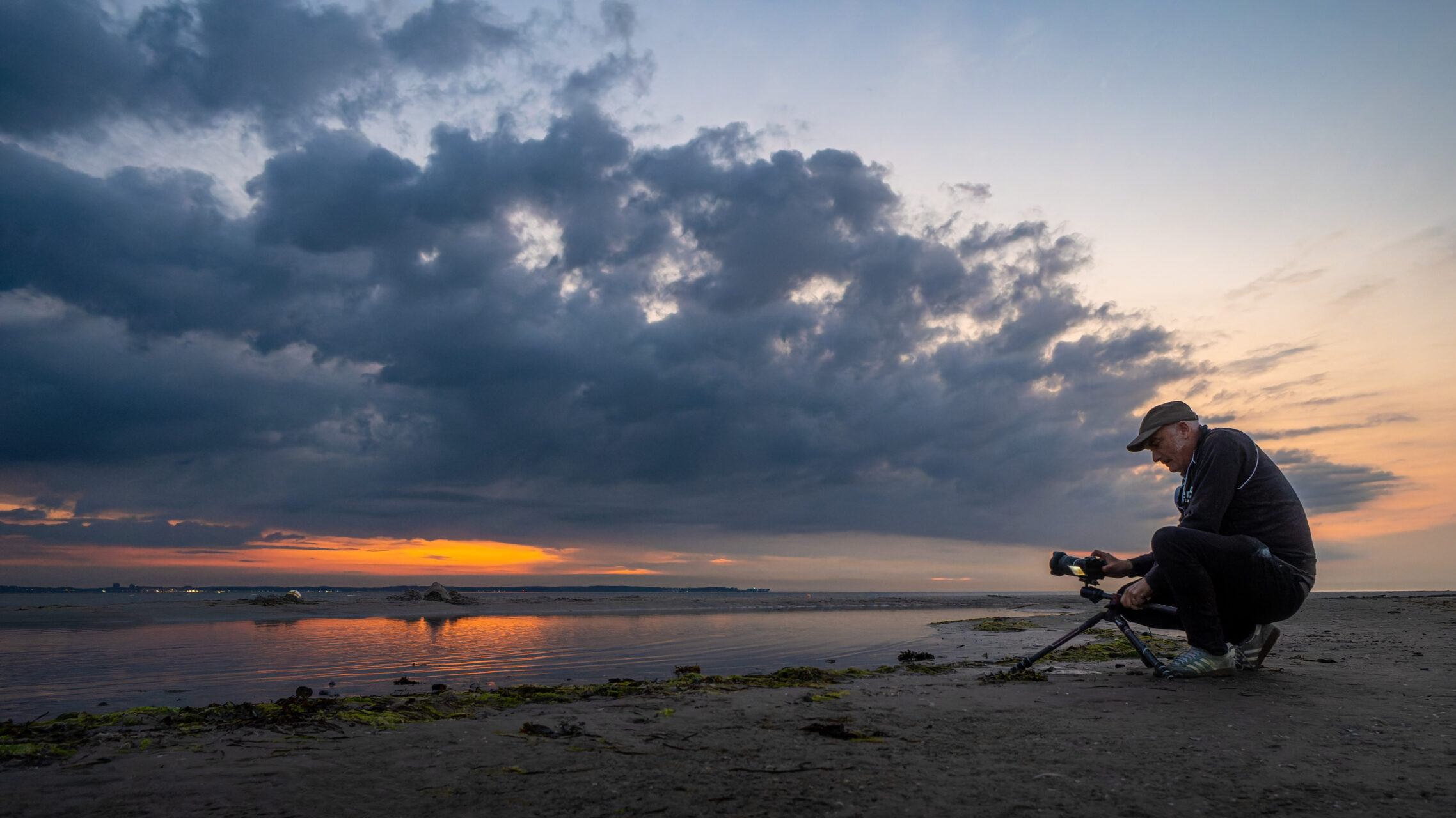 Ein Fotograf sitzt am Strand und richtet seine Kamera auf den Sonnenuntergang. Die dramatische Wolkenlandschaft spiegelt sich im Wasser, während das Licht des Abendhimmels die Szene beleuchtet.
