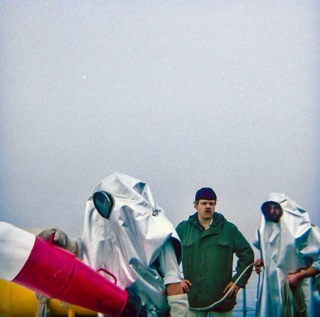 Three individuals in protective gear on a foggy marine environment, one man in a green jacket looks on while two others in silver suits prepare equipment. The scene reflects maritime safety practices and teamwork in challenging conditions.