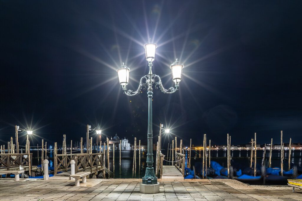 Beleuchteter Hafen mit Laterne bei Nacht in Venedig, Holzsteg und Boote im Hintergrund, ruhige Wasseroberfläche.