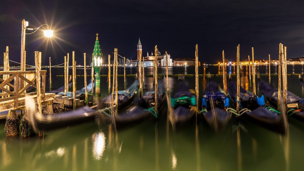 Die nächtliche Aussicht auf den venezianischen Hafen mit Gondeln, beleuchtetem Leuchtturm und historischen Gebäuden im Hintergrund, reflektiert im ruhigen Wasser.