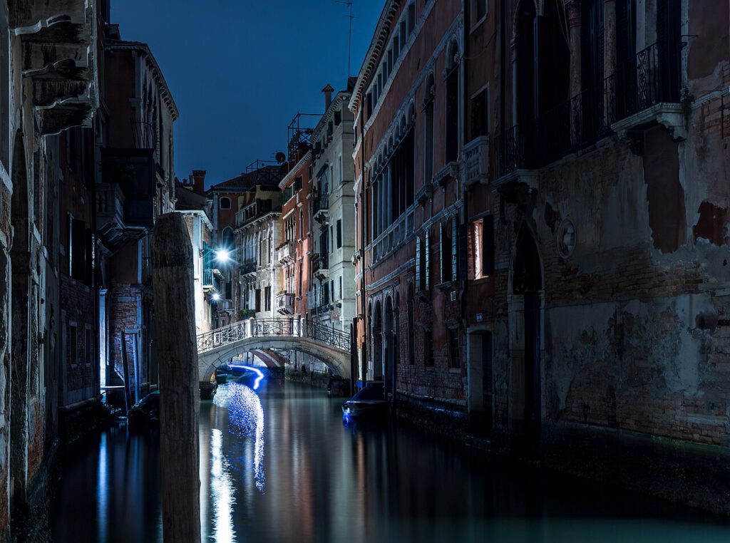 Venedig bei Nacht: ruhiger Kanal mit beleuchteter Brücke und historischen Gebäuden, reflektiert im Wasser. Ideal für romantische Spaziergänge und Fotografie.