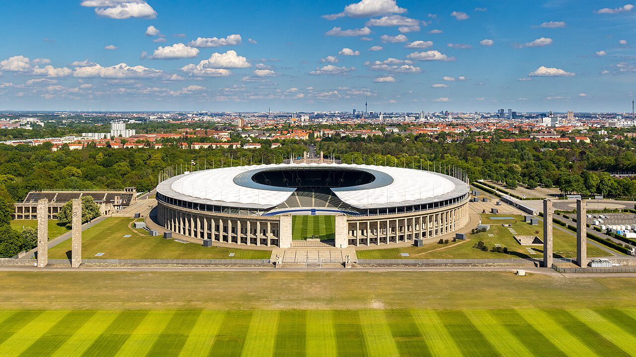 Panoramablick auf das Olympiastadion in Berlin, umgeben von grünen Rasenflächen und Bäumen, mit einer klaren blauen Himmel und Wolken im Hintergrund.