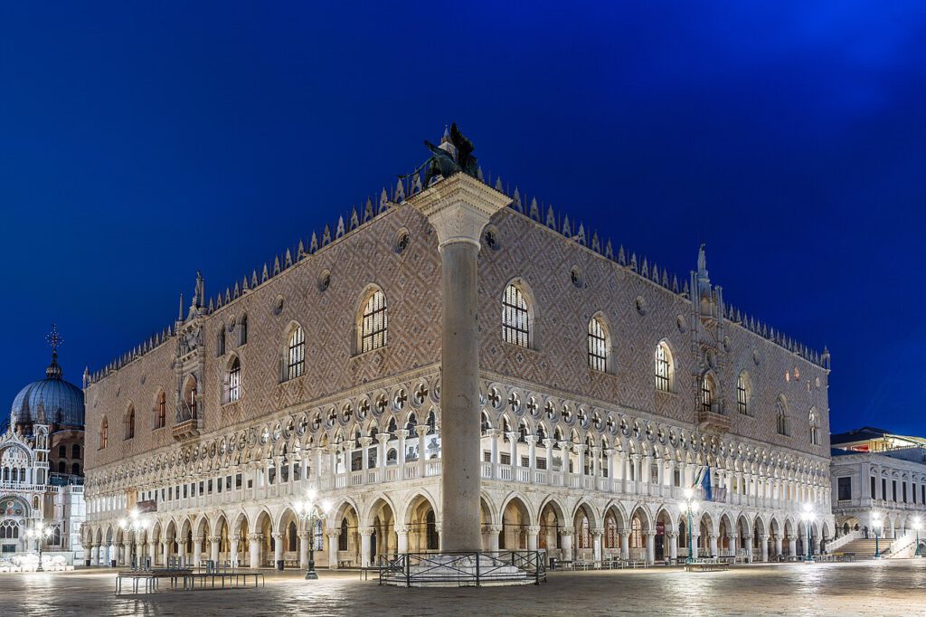 Historisches Gebäude in Venedig, Italien, bei Nacht. Das Bild zeigt den markanten Palazzo Ducale mit seiner charakteristischen Architektur und den Säulen im Vordergrund. Die sanften Lichter beleuchten die Fassade und schaffen eine malerische Atmosphäre.