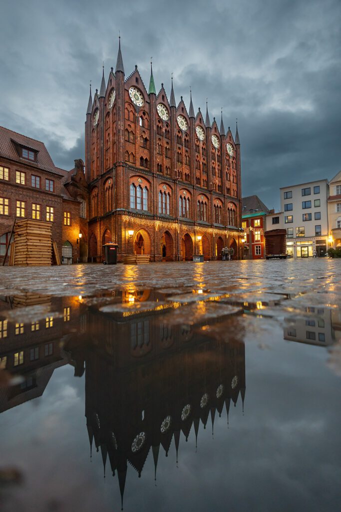 Historisches Gebäude im gotischen Stil mit markanten Türmen und Uhren, reflektiert in einer Pfütze auf dem Platz, umgeben von modernen und traditionellen Gebäuden unter einem bewölkten Himmel.