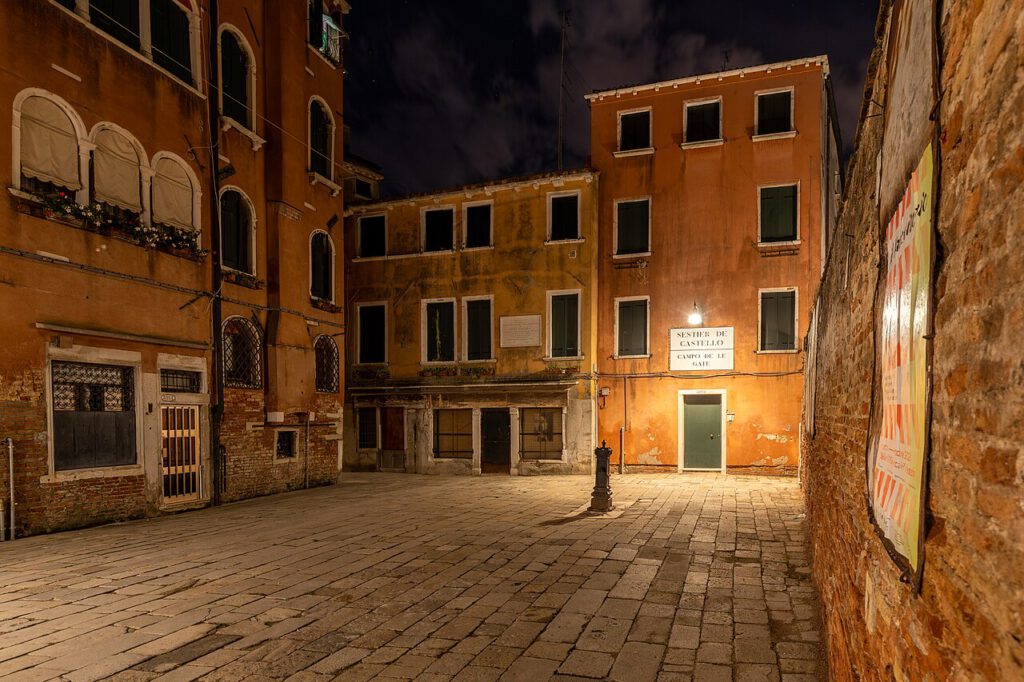 Nachtaufnahme eines ruhigen Platzes in Venedig mit historischen Gebäuden und einem beleuchteten Eingang. Die Wände sind in warmen Erdtönen gehalten, und der gepflasterte Boden reflektiert das Licht. Im Hintergrund ist ein Schild mit dem Schriftzug "SENTIER DE CASTELLO" zu sehen.