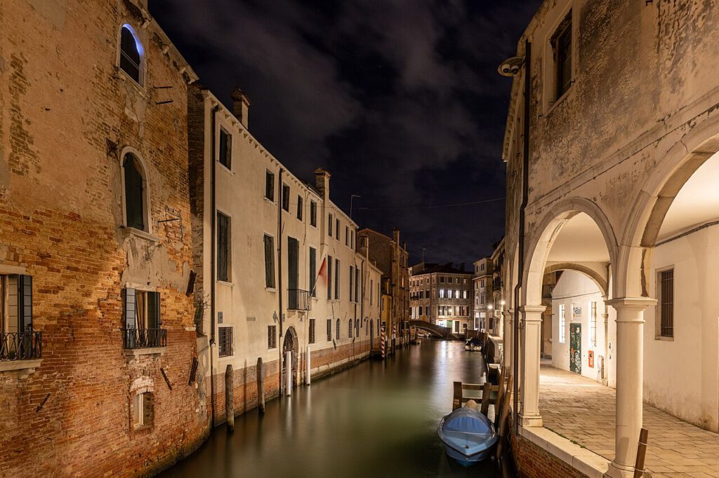 Nachtaufnahme eines venezianischen Kanals mit historischen Gebäuden, beleuchtetem Wasser und einem kleinen Boot, das am Steg festgemacht ist. Die Architektur zeigt typische Elemente Venedigs, während der Himmel mit Wolken bedeckt ist.