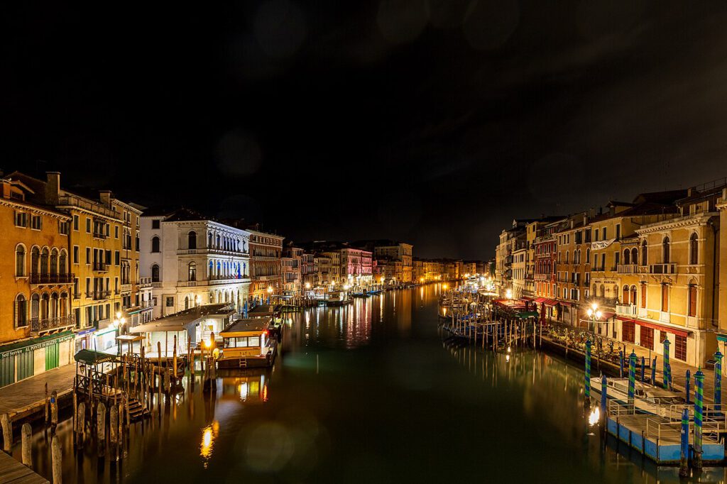 Venedig bei Nacht mit beleuchteten Gebäuden entlang des Kanals, reflektierenden Lichtern auf dem Wasser und Holzstegen. Ideal für Reisende, die die romantische Atmosphäre der Stadt erleben möchten.