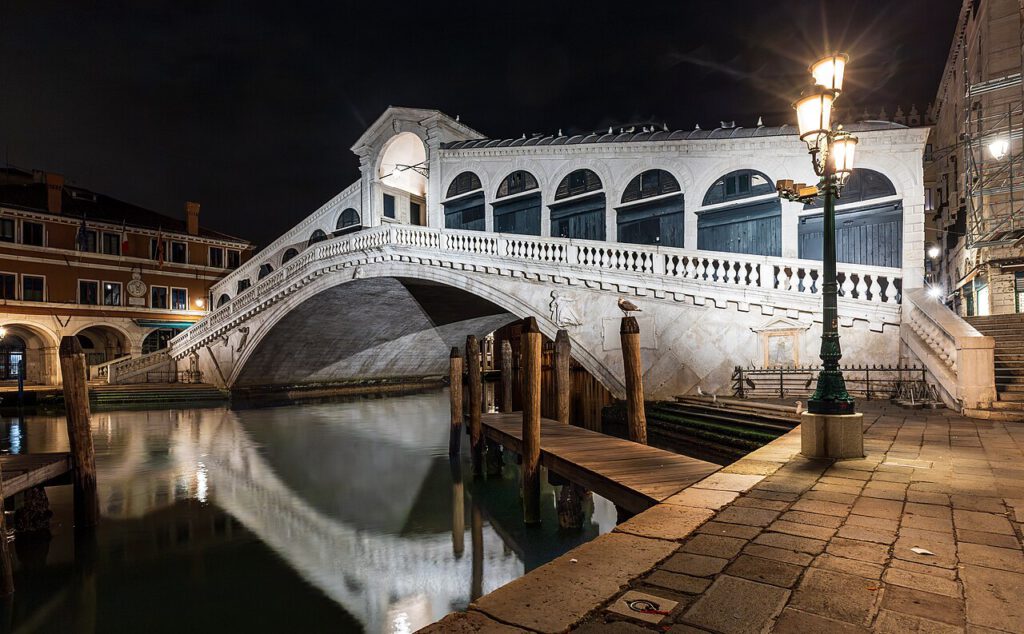 Rialtobrücke in Venedig bei Nacht, beleuchtet und reflektiert im Wasser, umgeben von historischen Gebäuden und Laternen.