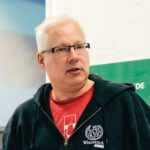 Man with short white hair and glasses speaking during an event, wearing a black hoodie over a red shirt with a graphic design.