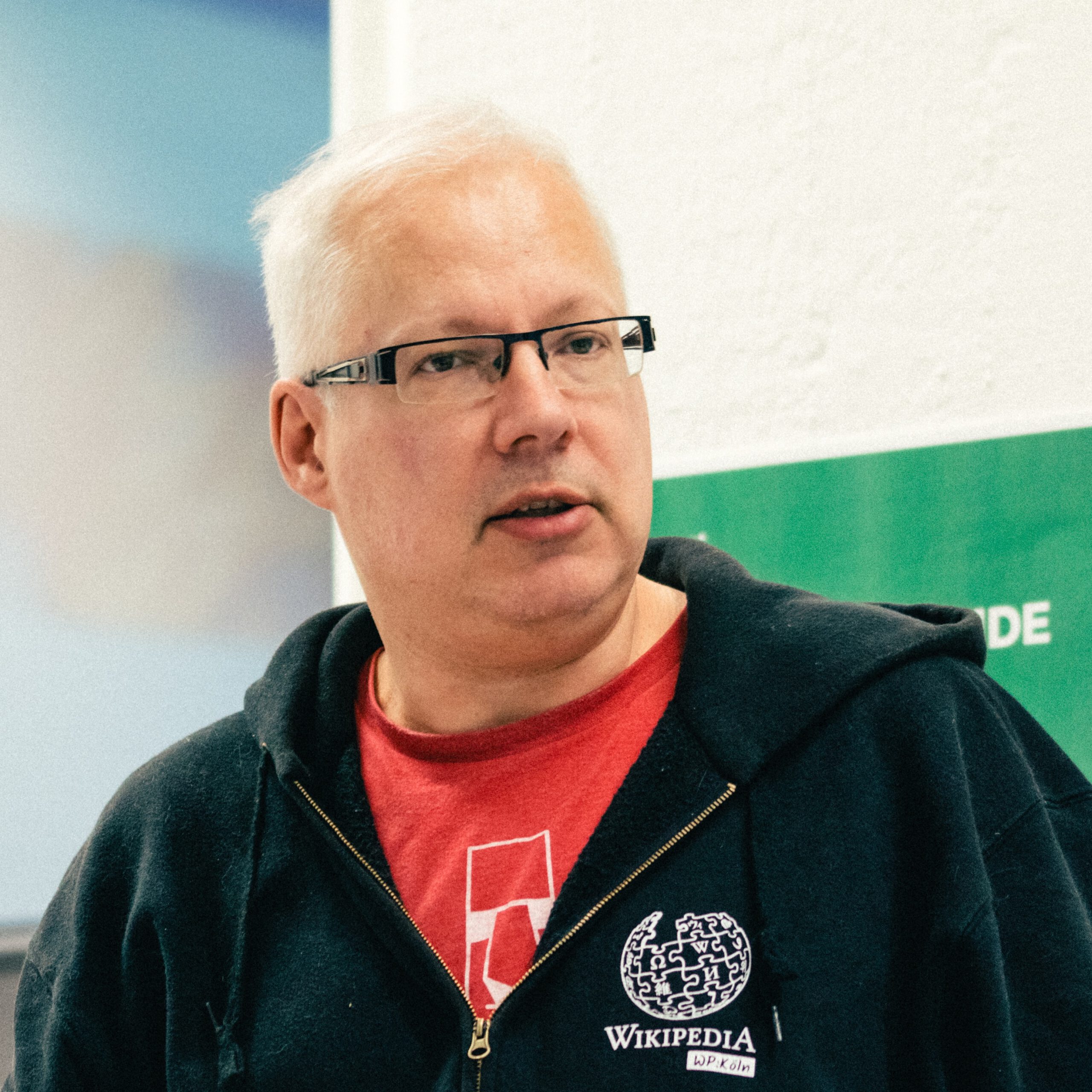 Image of a person with short, white hair, wearing glasses and a black hoodie with a Wikipedia logo, speaking attentively in a casual setting. The background features a green wall with text.