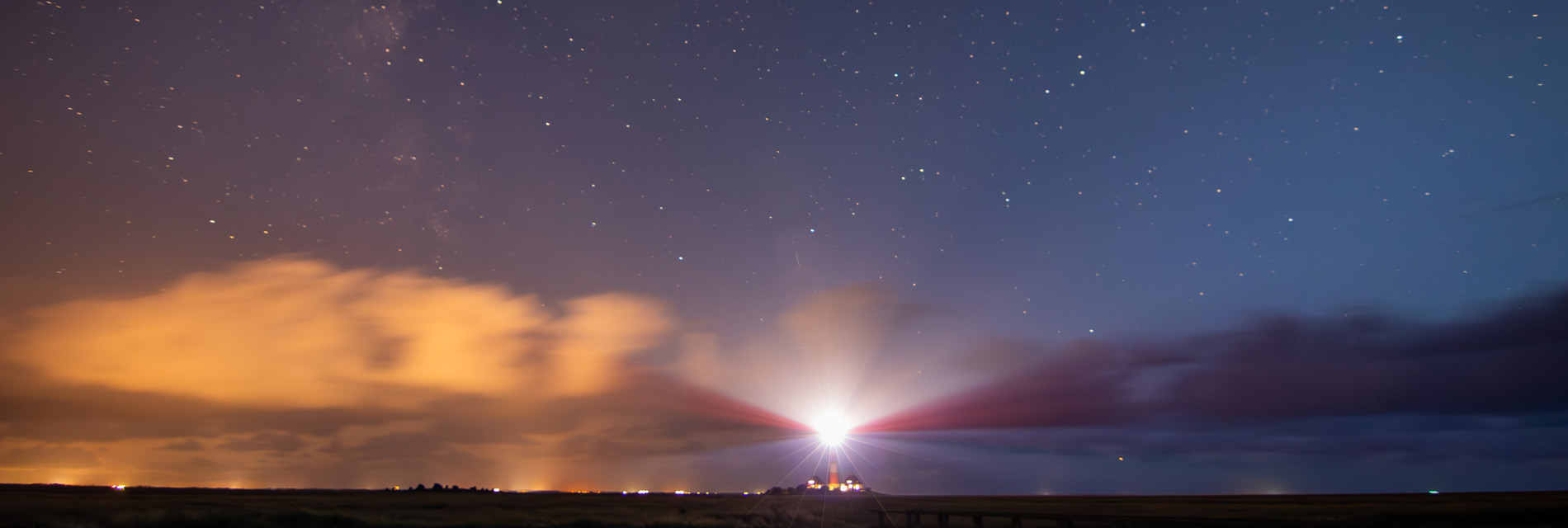 Leuchtturm bei Nacht, umgeben von einem klaren Sternenhimmel und sanften Wolken, die in warmen Farben leuchten. Die Lichtstrahlen des Leuchtturms durchbrechen die Dunkelheit und schaffen eine eindrucksvolle Atmosphäre. Ideal für Natur- und Landschaftsphotographie.