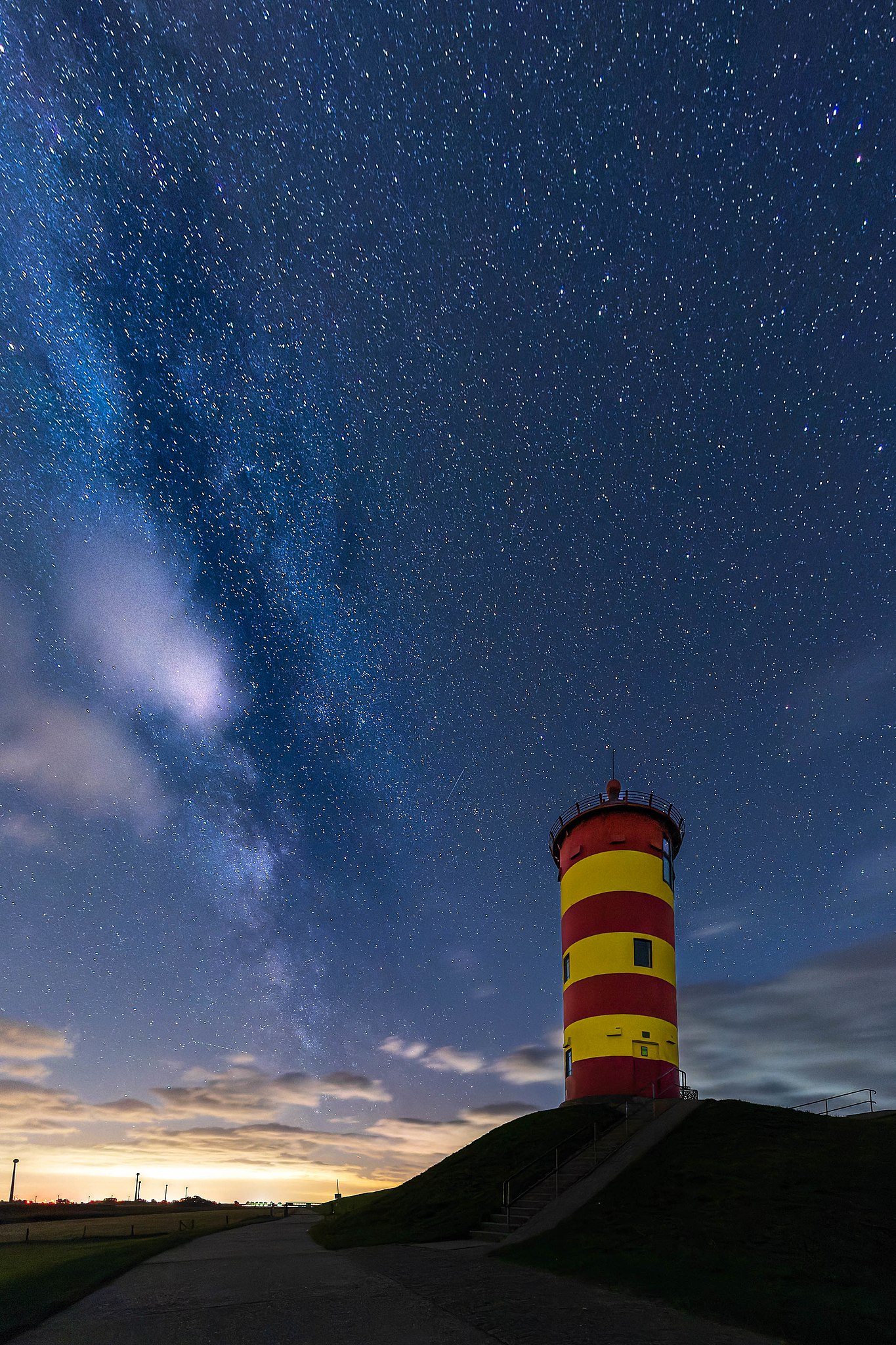Leuchtturm mit rot-gelber Streifenmuster unter einem sternenklaren Nachthimmel, umgeben von Wolken und dem Licht des Sonnenuntergangs. Ideal für Astronomie- und Naturfotografie.