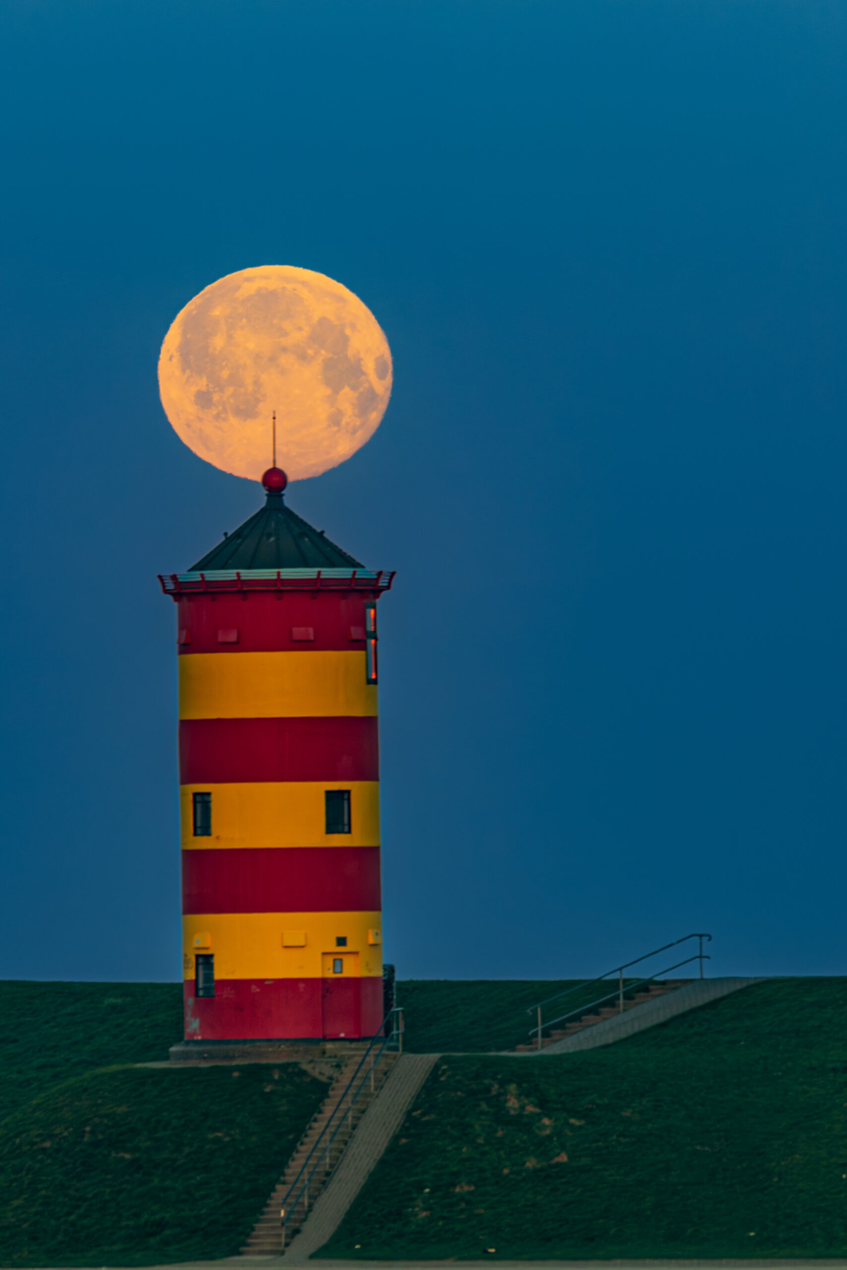 Leuchtturm mit rot-gelber Streifenfarbe vor einem klaren Abendhimmel, während der volle Mond direkt darüber aufgeht. Ideal für Landschafts- und Nachtfotografie.
