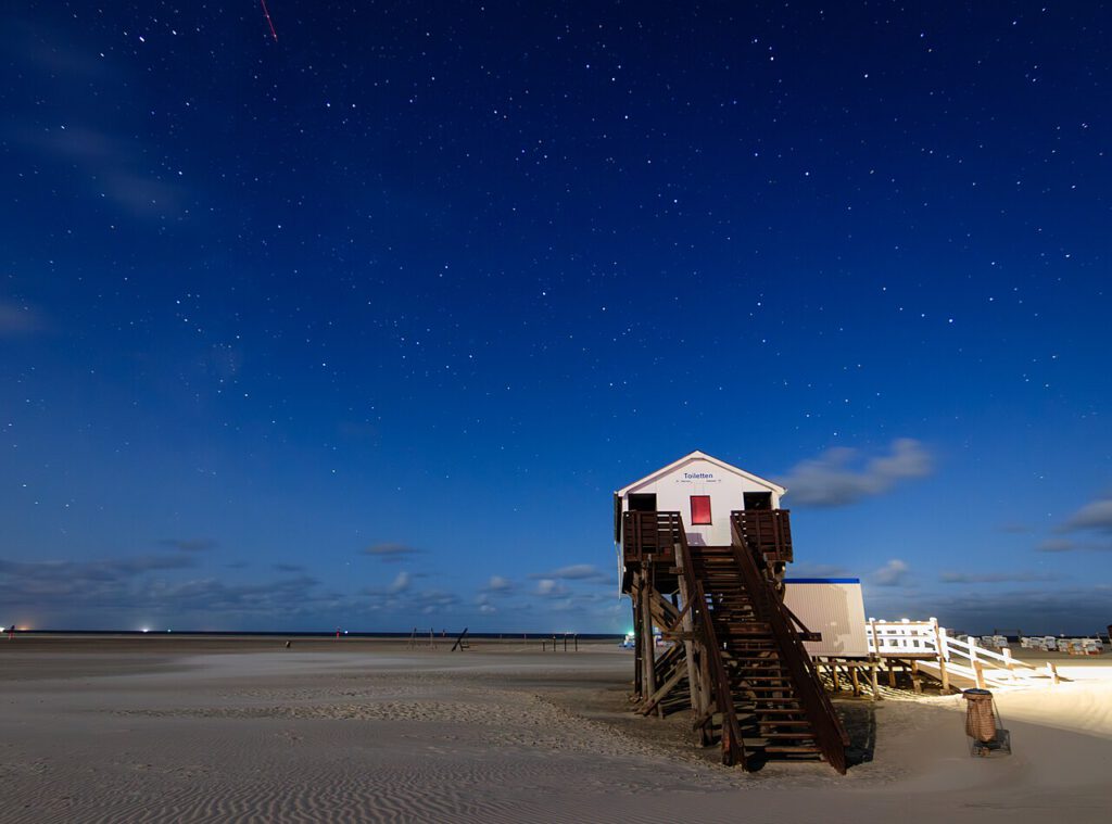 Ein beleuchtetes Toilettenhäuschen am Strand unter einem klaren Nachthimmel mit vielen Sternen. Die Sandfläche ist weitläufig und bietet einen ruhigen Ausblick auf das Meer.