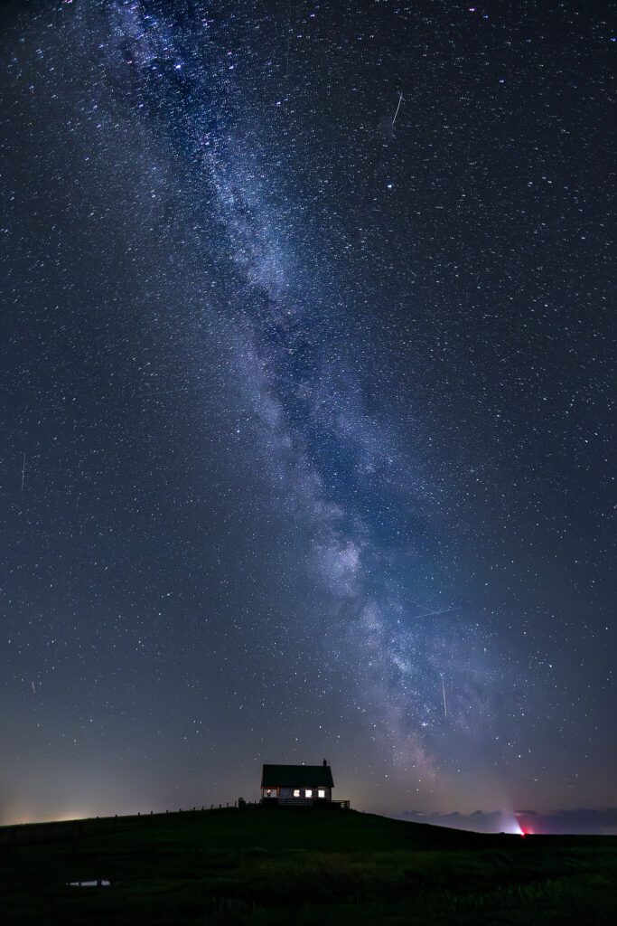 Sternenklare Nacht über einem einsamen Haus auf einem Hügel, mit der Milchstraße, die den Himmel durchzieht und mehreren Sternschnuppen, die sichtbar sind.