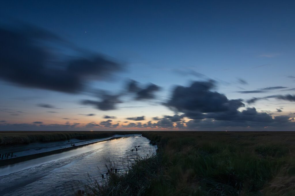 Sonnenuntergang über einem ruhigen Gewässer, umgeben von sanften Hügeln und Wolken am Himmel, mit reflektierendem Licht auf der Wasseroberfläche.