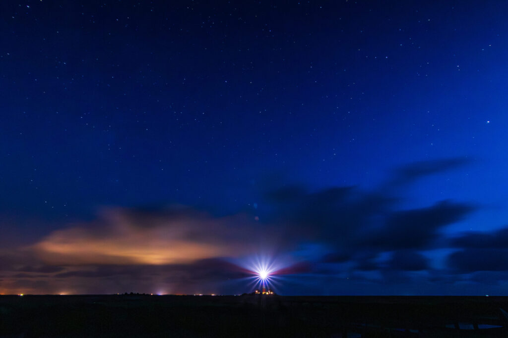 Leuchtturm im nächtlichen Himmel mit Sternen und Wolken, umgeben von sanften Lichtern am Horizont.