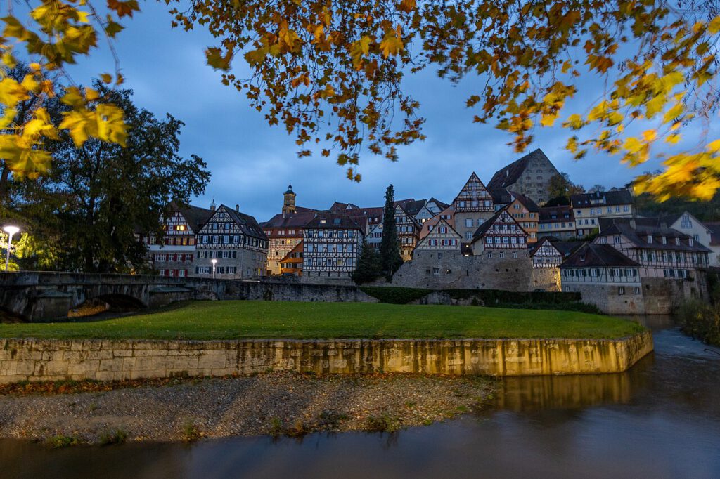 Historische Altstadt von Schwäbisch Hall am Abend, mit Fachwerkhäusern entlang des Flusses und buntem Herbstlaub im Vordergrund.