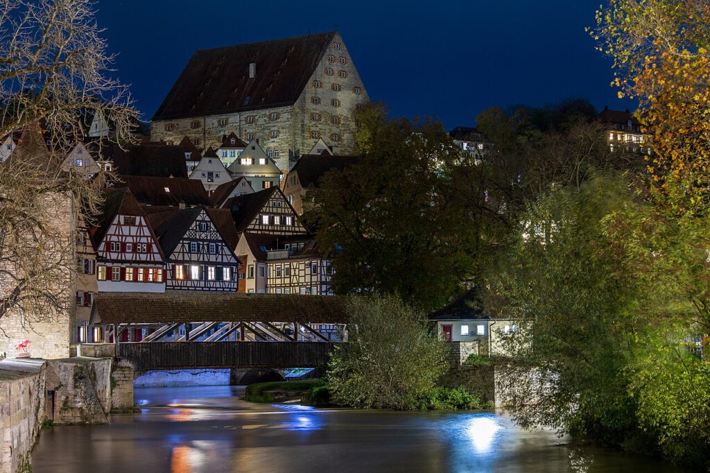 Historische Fachwerkhäuser entlang eines Flusses bei Nacht, mit beleuchteten Fenstern und einer Brücke im Vordergrund, vor der Kulisse einer alten Burg in Deutschland.