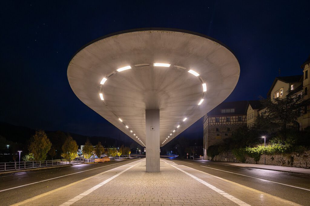 Moderne Überdachung über einer Straße bei Nacht, beleuchtet von LED-Lichtern, umgeben von Bäumen und historischen Gebäuden.