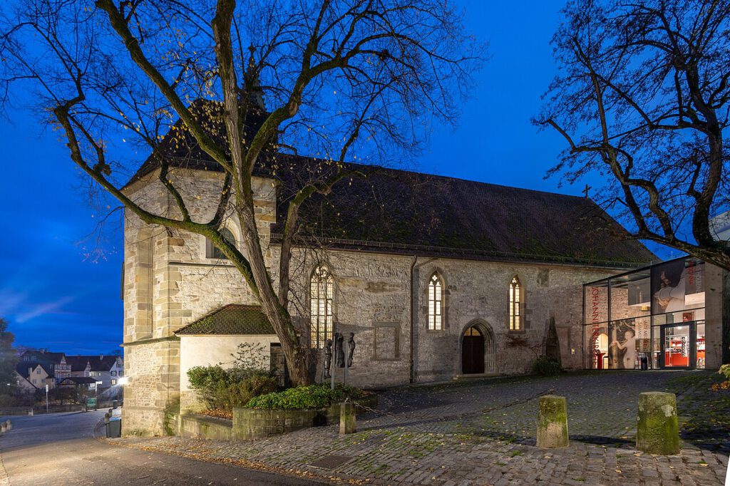 Historisches Gebäude bei Nacht, das eine steinerne Kirche mit gotischen Fenstern zeigt, umgeben von Bäumen und einem gepflasterten Platz. Im Hintergrund sind moderne Elemente sichtbar, die die Verbindung von Tradition und zeitgenössischer Architektur verdeutlichen.